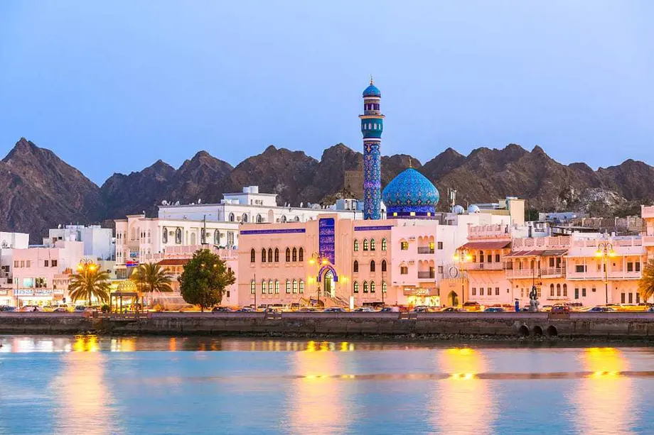 The Mutrah waterfront in Muscat at dusk, with a blue-domed mosque against the mountains
